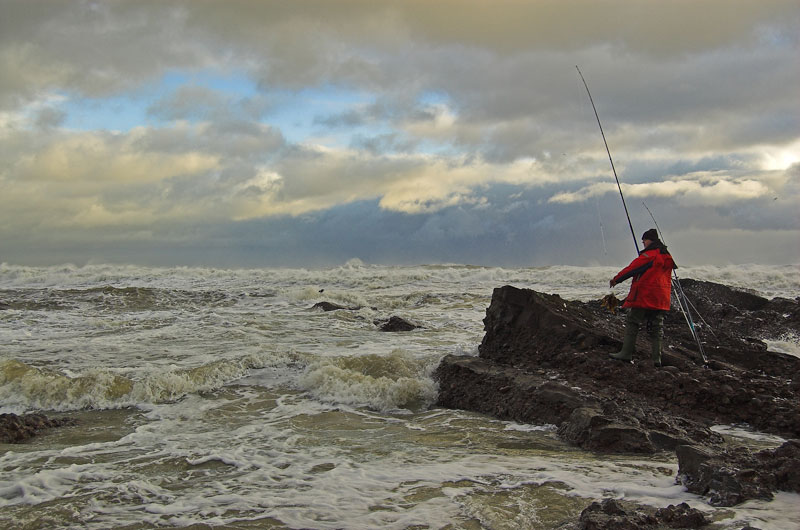 an angler on a rocky shore with churning seas